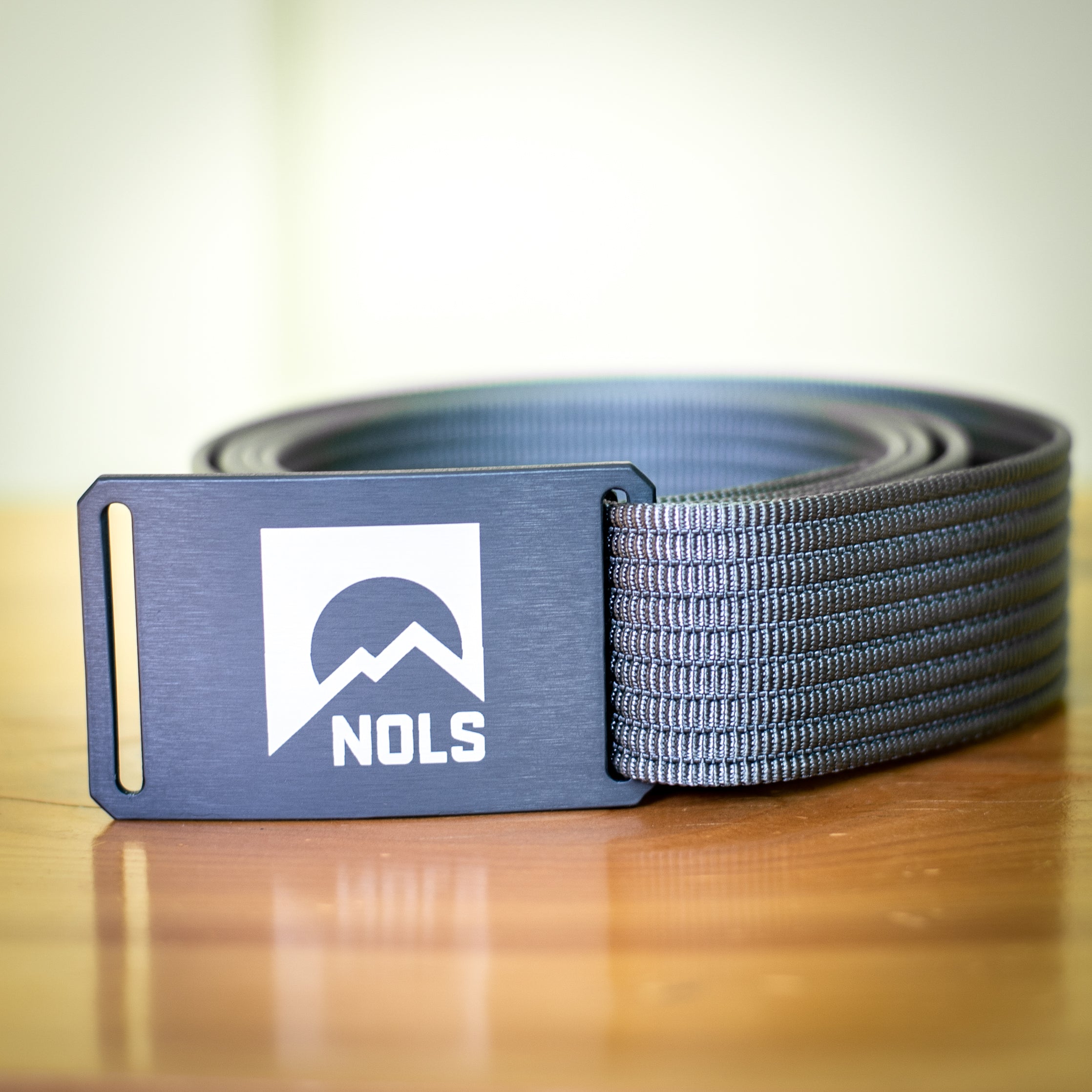 GRIP6 Belt with NOLS Logo featuring a gunmetal aluminum buckle with white mountain logo design, paired with durable black nylon webbing strap, displayed on wooden surface