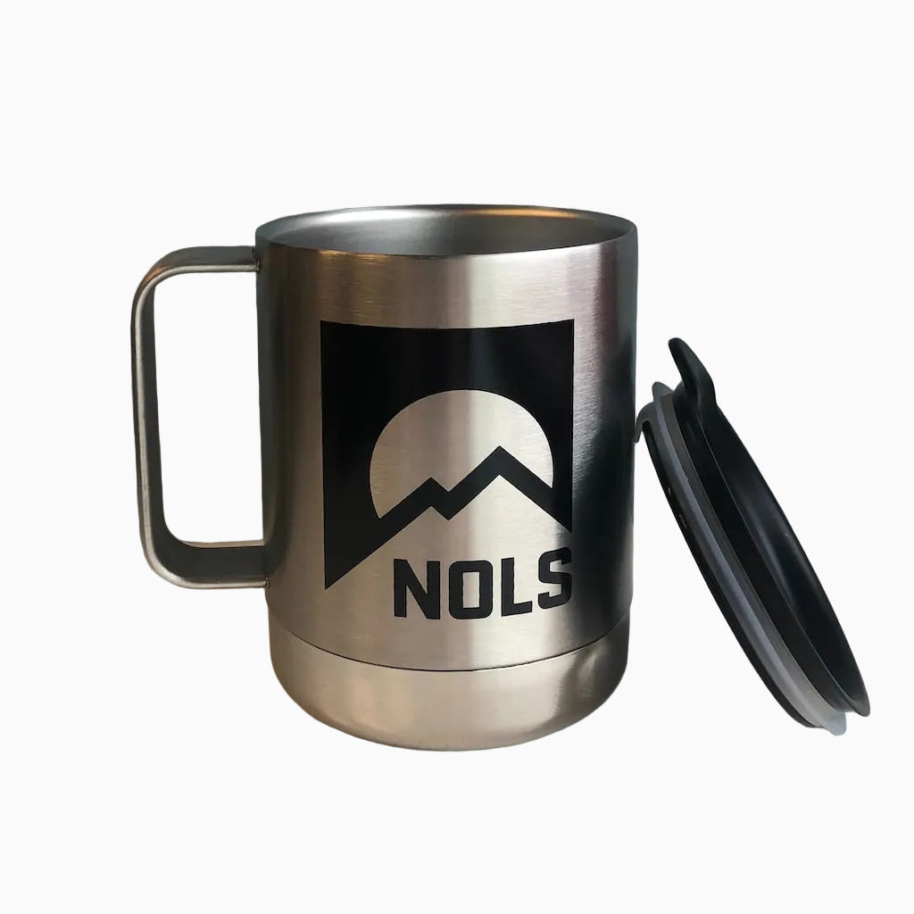 Klean Kanteen NOLS Camp Mug in brushed stainless steel with black NOLS mountain logo, featuring insulated double-wall construction and removable black splash-proof lid, displayed on white surface against gray wall