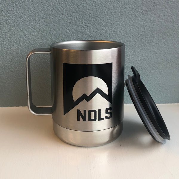 Klean Kanteen NOLS Camp Mug in brushed stainless steel with black NOLS mountain logo, featuring insulated double-wall construction and removable black splash-proof lid, displayed on white surface against gray wall