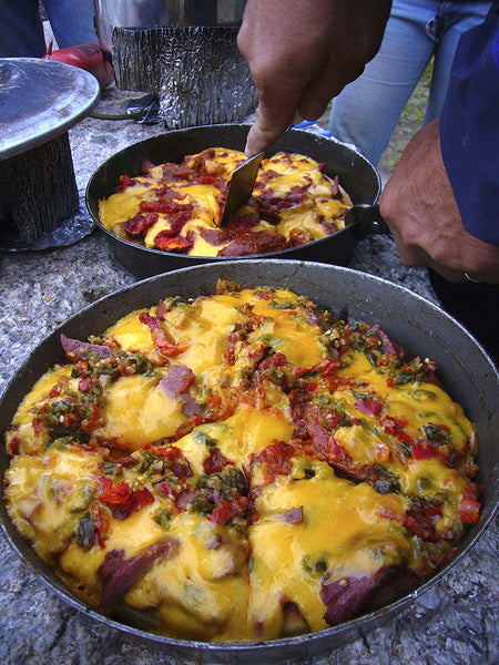 Expedition Fry-Bake pan filled with cheesy mountain breakfast loaded with melted cheese, bacon, and vegetables, being served outdoors on a stone surface, demonstrating the pan's versatile cooking capabilities in wilderness settings.