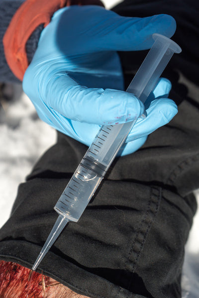 Irrigation Syringe 12cc held in blue-gloved hand, demonstrating clear plastic medical syringe with measurement markings for precise wound cleaning and debris removal in wilderness first aid situations